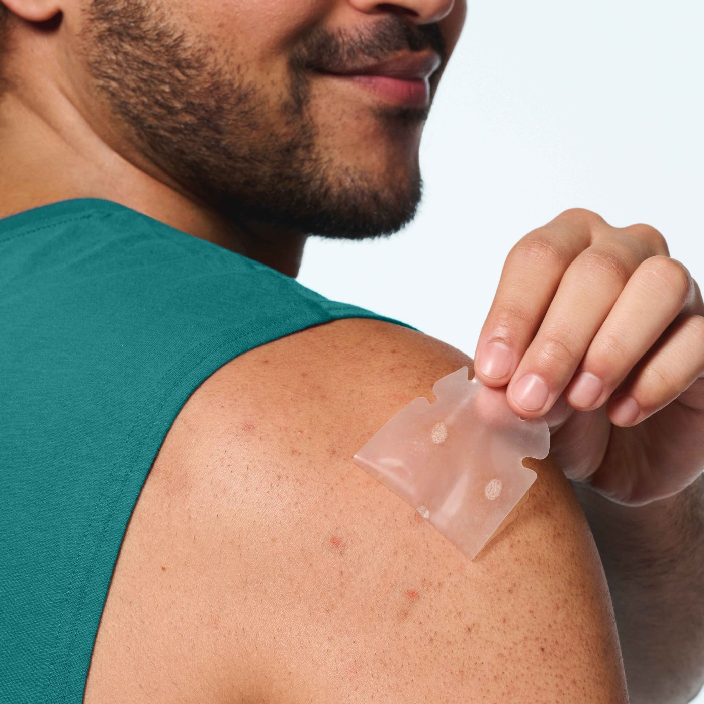 Man in teal tank top lifting a large clear hydrocolloid Mighty Patch off his shoulder, two white pimple spots visible on the film after eight hours of wear